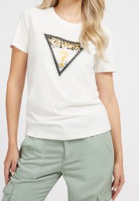 Camiseta de algodón blanca de manga corta, con un gráfico triangular en negro y plateado que presenta el texto "GUESS" y un signo de interrogación en dorado.