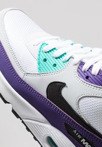 Baskets blanches avec accents violets et bleus canard, tige en synthétique texturé, sections en mesh, logo noir et amorti à air visible dans la semelle.