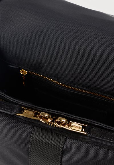 Μαύρη υφασμάτινη τσάντα με δύο χρυσά φερμουάρ με την επιγραφή «Moschino» και ένα μερικώς ορατό χρυσό φερμουάρ μέσα στο ανοιχτό διαμέρισμα.