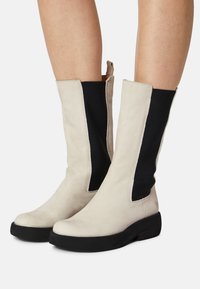 Kozaki do kolan z miękkiej off-white skóry z czarnymi elastycznymi panelami po bokach, okrągłym noskiem i grubą czarną podeszwą. Minimalistyczny design.