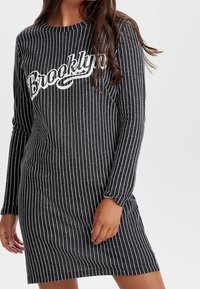 Pitkähihaisen, harmaaraidallisen mekon etuosassa on valkoinen teksti "Brooklyn". Kangas näyttää pehmeältä, jossa on ribbikuviointi ja rento istuvuus.