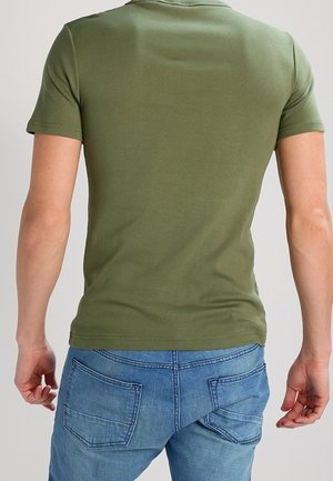 Homme portant un t-shirt vert olive à manches courtes et un jean bleu en denim, vu de dos sur un fond uni.