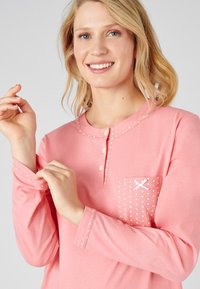 Maglia rosa chiaro a maniche lunghe con collo henley abbottonato, pois bianchi sulla tasca e cuciture decorative sulle maniche.