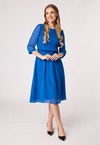 Blauwe chiffon jurk met een geplooide buste, driekwart mouwen en een gestrikte tailleband, gecombineerd met zwarte puntige hakken.