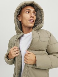 Beige pufferjakke med hætte, med quiltet tekstur, lynlås-lukning og mærke-logo på brystet, båret over en hvid skjorte.