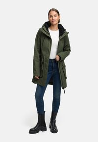 Frau, die eine grüne Parka mit Kapuze und schwarzem Fleece-Futter, ein weißes Oberteil, blaue enge Jeans und schwarze klobige Stiefel trägt, vor einem weißen Hintergrund steht.
