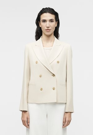 Γυναίκα με σκούρα μαλλιά που φοράει κρεμ σακάκι με διπλό πέτο και άσπρο παντελόνι, όρθια μπροστά σε απλό φωτεινό φόντο.