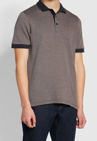 Homme portant un polo à manches courtes noir et blanc à motifs, avec un col et des poignets noirs, associé à un jean bleu foncé.