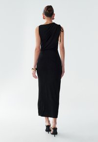 Kvinde med hår i en knold bærer en ærmeløs sort midi-kjole med ruching på den ene skulder, sorte højhælede sko, et guldbelagt armbånd og runde øreringe.