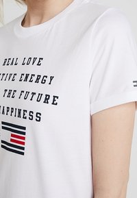 Hvid T-shirt med sort tekst, der står "Ægte Kærlighed", "Positiv Energi", "Fremtiden", "Lykke" og et sort og rødt stribet grafisk mønster nedenfor.