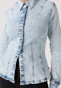Γυναίκα που φοράει εφαρμοστή τζιν ζακέτα σε ανοιχτό γαλάζιο χρώμα με όξινο πλύσιμο, με κουμπιά μπροστά και μυτερό γιακά, συνδυασμένη με μαύρο παντελόνι.
