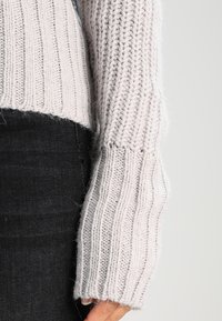 Maglione lavorato a maglia grigio chiaro con righe verticali, ampie maniche e un design cropped. Indossato sopra jeans scuri, evidenziando la texture e il materiale.