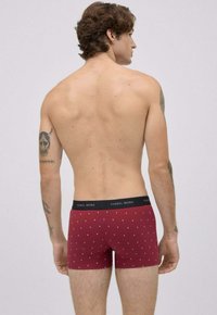 Hombre con tatuajes en los brazos, vistiendo calzoncillos tipo bóxer de color burdeos con un diseño y cinturilla negra etiquetada "Ysabel Mora," de espaldas contra un fondo liso.