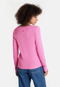 Frau mit lockigem Haar, die ein langärmliges pinkes Shirt und eine blaue Jeans trägt, mit dem Rücken zur Kamera und nach rechts schauend, vor einem weißen Hintergrund.