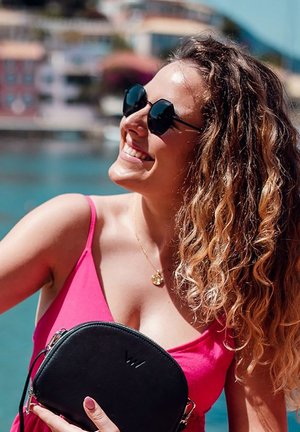 Černá kulatá kabelka se zipem, držena v jedné ruce. Taška má lesklou texturu s logem. V pozadí je modrá voda a květinový výhled.