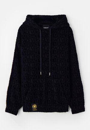 Must räpane toest, millel on sametine tekstuur, kogu pinnale kantud Versace logo mustriga, reguleeritavad nöörid ja kollane logo silt allääres.