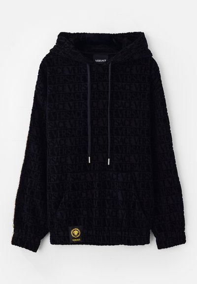 Juodas džemperis su aksominiu tekstūru, pasižymintis viso paviršiaus Versace logotipo raštu, reguliuojamomis raišteliais ir geltonu logotipo užtrauktuku ties apačia.