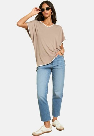 Beige V-Ausschnitt-T-Shirt mit weißen Akzenten, lockerer Schnitt, kombiniert mit hellblauen geraden Jeans und weißen Sneakern. Model trägt runde schwarze Sonnenbrille.