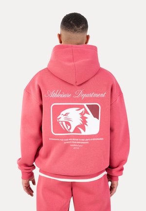 Mann in rosa Kapuzenpullover und Hose, von hinten gesehen, auf dem Kapuzenpullover ein weißes Logo einer brüllenden Panther mit dem Schriftzug "Athleisure Department" darüber.