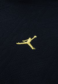 Čierna textúrovaná tkanina s výrazným žltým vyšívaným logom Jumpman na ľavej strane, ktorá zdôrazňuje športový dizajn.