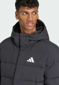 Crna podstavljena jakna s visokim ovratnikom, s bijelim logom na prsima. Glatka tekstura s modernim, strukturiranim dizajnom.