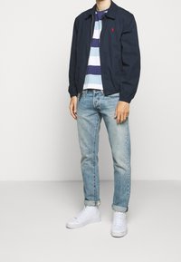 Giacca leggera navy con colletto alto, abbinata a una maglietta a righe blu e bianca, jeans in denim chiaro e sneakers bianche.