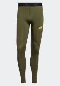 Oliwkowe legginsy sportowe wykonane z gładkiego, elastycznego materiału. Posiadają czarny elastyczny pas z logo Adidasa w kolorze żółtym.