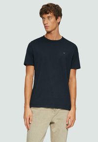 T-shirt in cotone blu navy a maniche corte con colletto tondo e un piccolo logo sul lato sinistro del petto, abbinato a pantaloni beige chiaro.