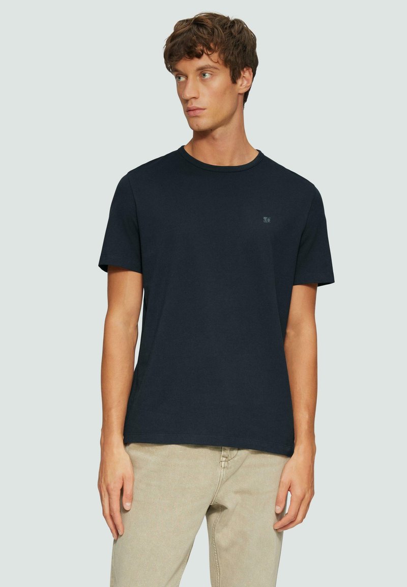 T-shirt in cotone blu navy a maniche corte con colletto tondo e un piccolo logo sul lato sinistro del petto, abbinato a pantaloni beige chiaro.