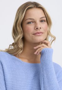 Jeune femme aux cheveux blonds ondulés portant un pull en maille bleu clair, reposant son menton sur sa main devant un fond blanc.