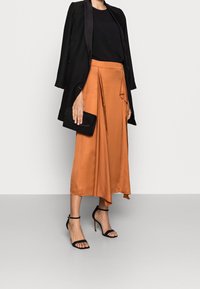 Malli, joka on pukeutunut mustaan bleiseriin ja puseroon, yhdistettynä oranssiin satiinihameeseen ja mustiin remmikenkiin, pitää mustaa clutch-laukkua. Neutraali tausta.