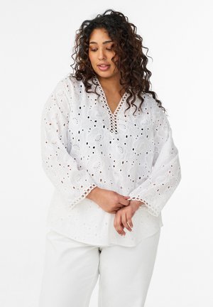 Käärus juustega naine, kes kannab valget tikandiga pika varrukaga pluusi ja valgeid pükse, poseerib käed ees kokku pandud.