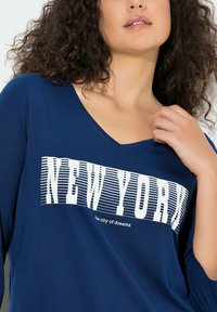 Marineblaues Langarm-T-Shirt mit V-Ausschnitt, versehen mit weißem "NEW YORK"-Schriftzug und gestreiften Details. Weicher Stoff und entspannte Passform.