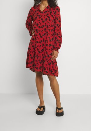 Robe rouge avec un motif floral noir, manches longues, décolleté en V et jupe à volants. Portée avec des sandales noires à semelle épaisse.