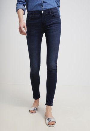 Person iført mørke skinny jeans, en opsmøget denimskjorte og sølvfarvede åbne slides stående på et lyst gulv.