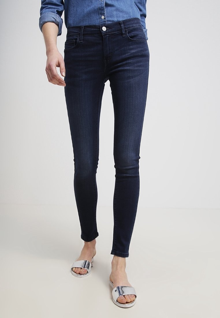 Personne portant un jean skinny foncé, une chemise en denim retroussée et des sandales argentées à bout ouvert, debout sur un sol clair.