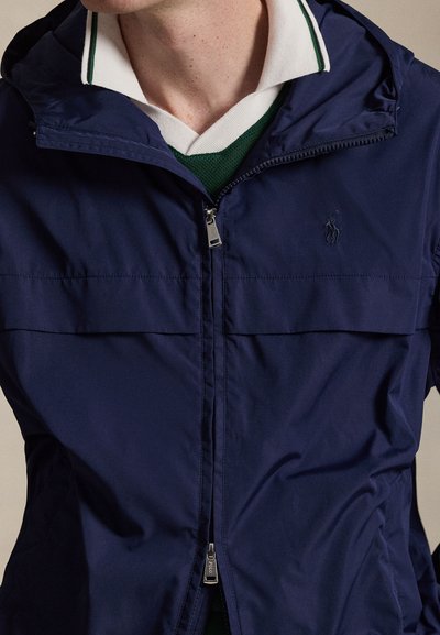 Homme portant une veste zippée bleu marine superposée à une chemise blanche à col avec des bordures vertes et un pull vert.