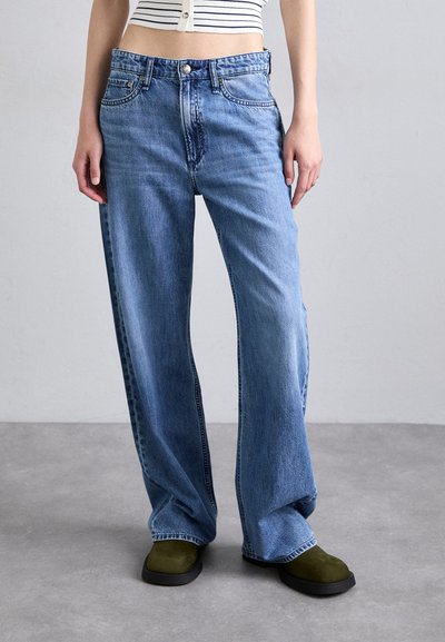 Jeans en denim bleu clair à jambes larges, avec une taille haute, cinq poches et une fermeture à bouton, associés à des chaussures slip-on olive.