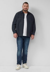 Donkerblauwe button-up shirt over een wit T-shirt, gecombineerd met blauwe jeans en lichte sneakers. Heeft twee voorzakken en lange mouwen.