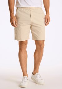 Shorts beiges avec une taille élastique, fermeture à bouton et poches latérales, fabriqués en tissu léger, avec une texture lisse et une coupe ajustée.