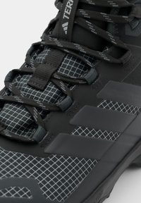 Close-up of black Adidas outdoor shoe with grid pattern, black reflective laces, and three matte stripes on the side.