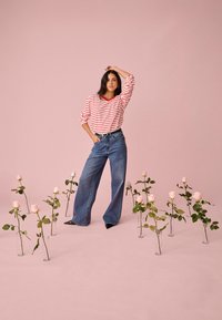 Naine seisab käsi peas, seljas punaste triipudega särk ja laiad teksad, ümbritsetud klaasvaasides roosadest roosidest roosal põrandal ja seinal.