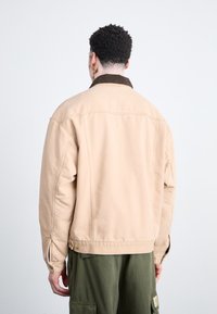 Chaqueta de algodón color caqui con un cuello de pana marrón, presenta botones en los puños y un ajuste relajado. Superficie texturizada con costuras visibles.