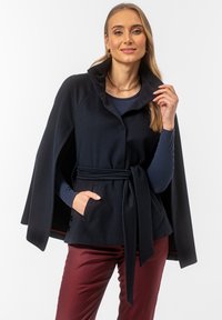 Granatowa kurtka w stylu peleryny z miękkiego materiału, z wysokim kołnierzem, paskiem w talii i bocznymi kieszeniami. Nałożona na granatową bluzkę i bordowe spodnie.