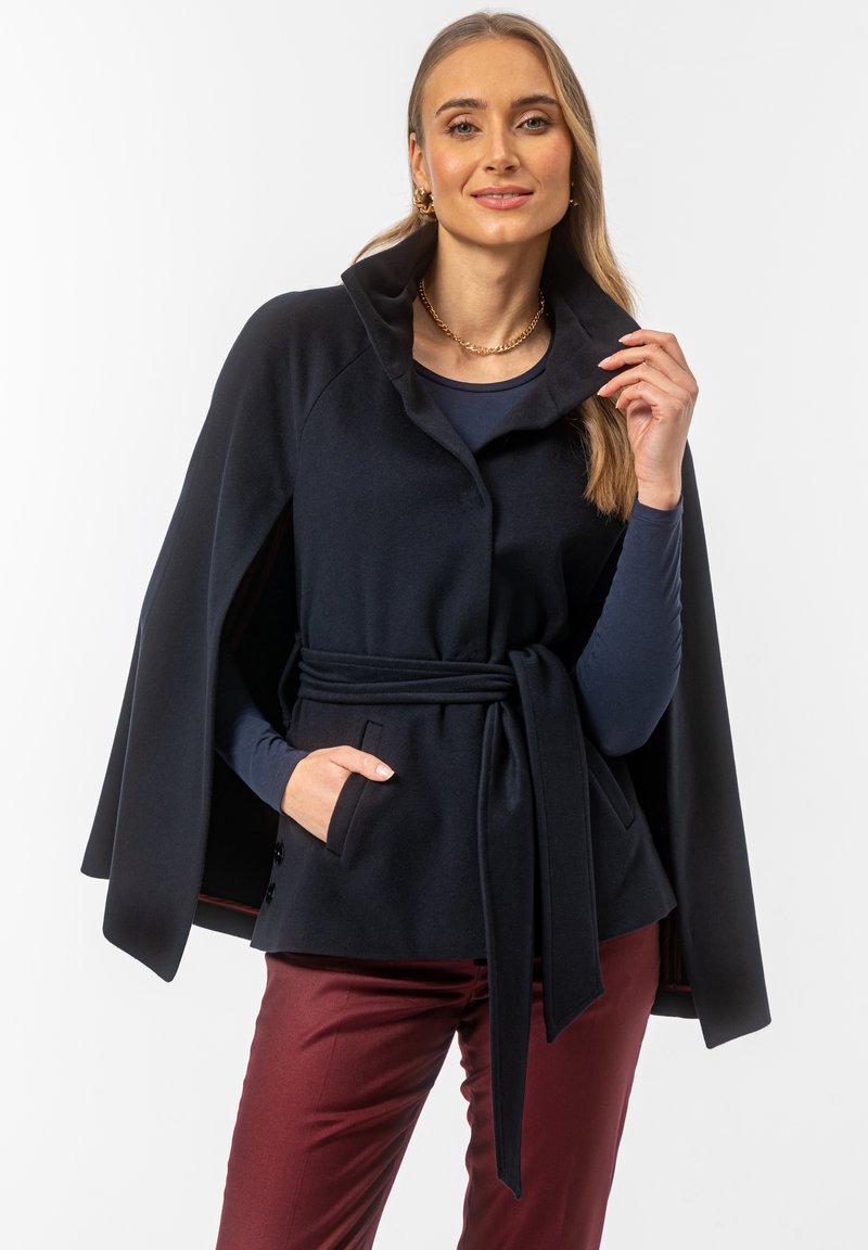 Granatowa kurtka w stylu peleryny z miękkiego materiału, z wysokim kołnierzem, paskiem w talii i bocznymi kieszeniami. Nałożona na granatową bluzkę i bordowe spodnie.