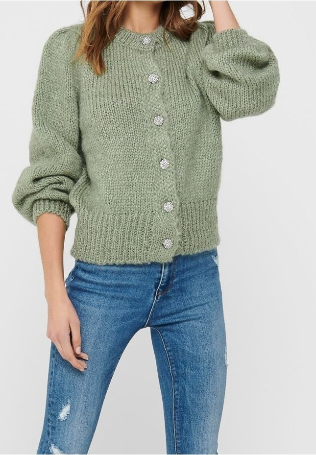 Zielony sweter robiony na drutach z przerośniętymi rękawami, sześcioma dekoracyjnymi guzikami i ściągaczem na dole; noszony na niebieskie, postrzępione dżinsy.