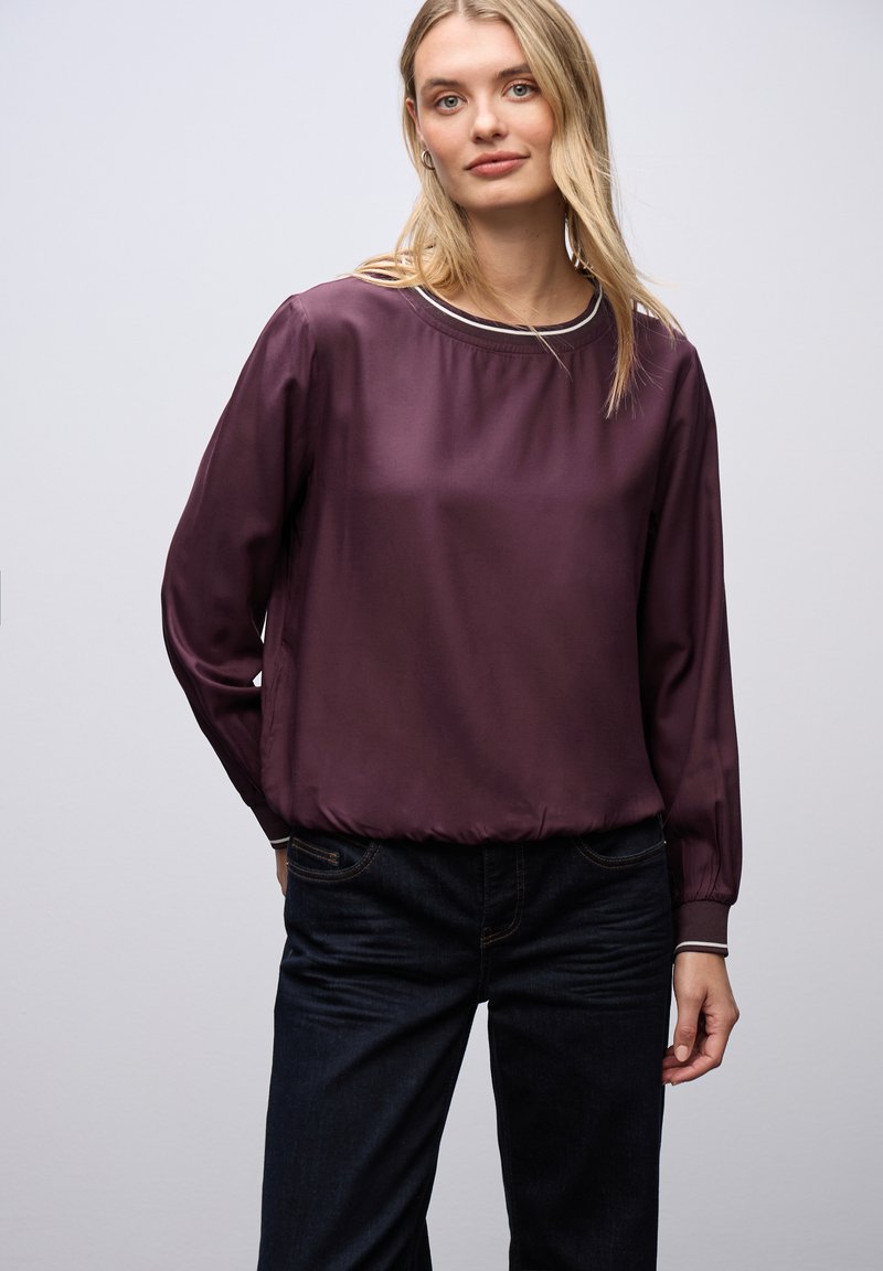 Blusa de satén de manga larga en un profundo color púrpura con cuello redondo y dobladillo elástico. Presenta un ribete en contraste alrededor del cuello y los puños.