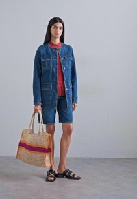 Jeansjacke mit Taschen, rotes Hemd, Jeansshorts, schwarze Sandalen und eine geflochtene Tragetasche mit einem mehrfarbigen Streifen.