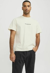 Cremfarbener T-Shirt mit kurzen Ärmeln featuring einem schwarzen grafischen Schriftzug auf der Brust, kombiniert mit dunkelgrauen, locker sitzenden Jeans. Einfaches und sauberes Design.