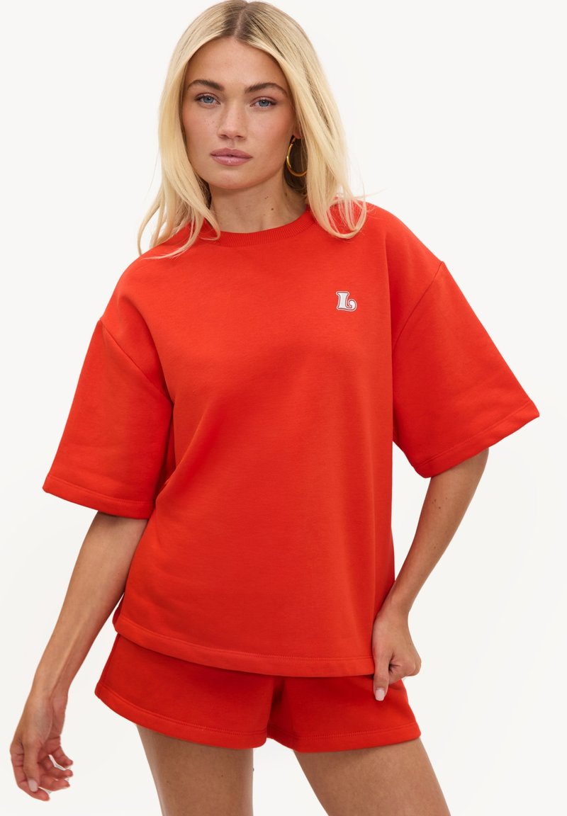 Femme blonde portant une chemise rouge vif à manches courtes oversize et un short assorti sur un fond uni.